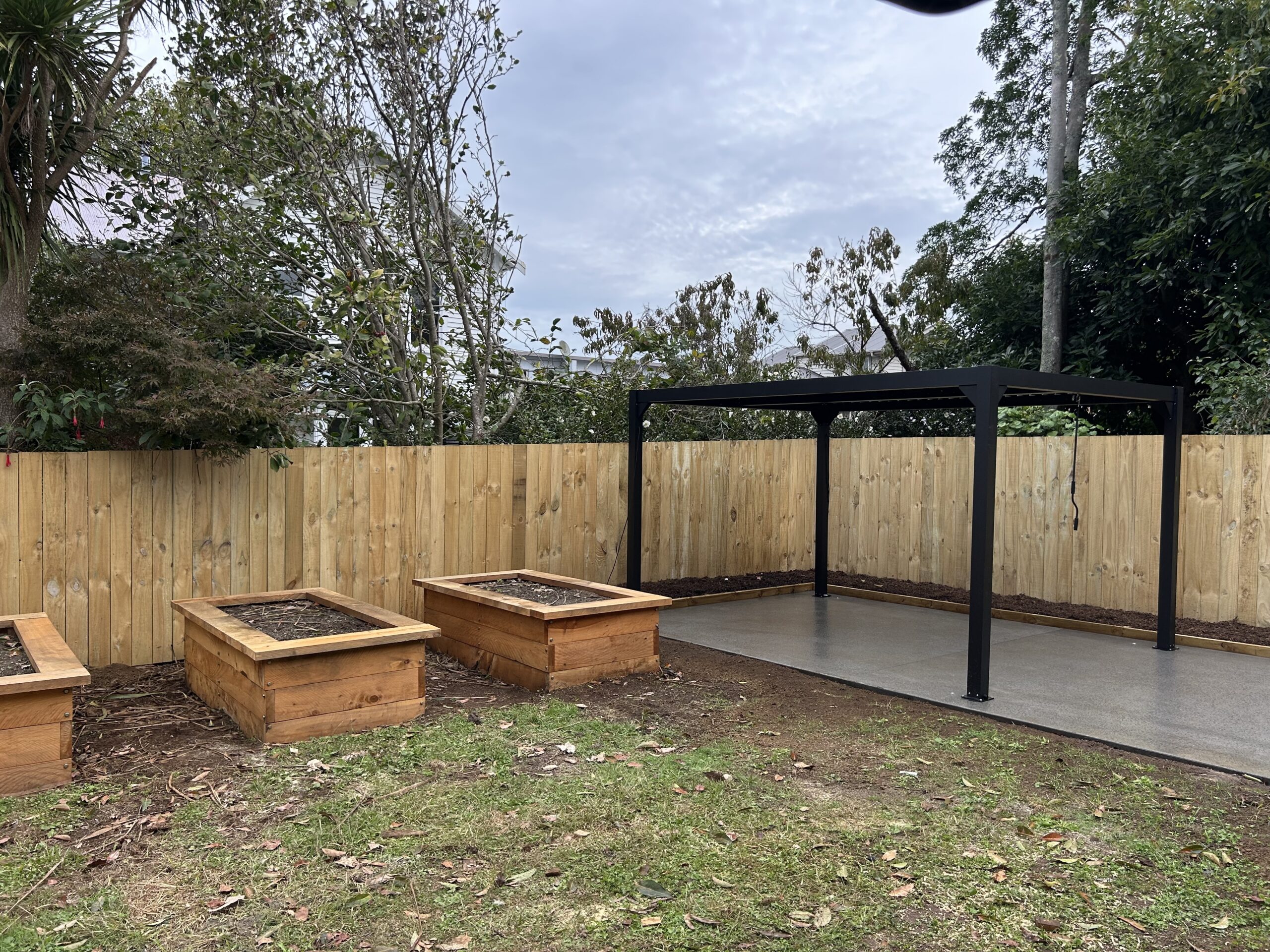 Outdoor living space with aluminium pergola, Macrocarpa raised garden beds, decorative concrete patio and new rough-sawn timber boundary fence in Auckland.